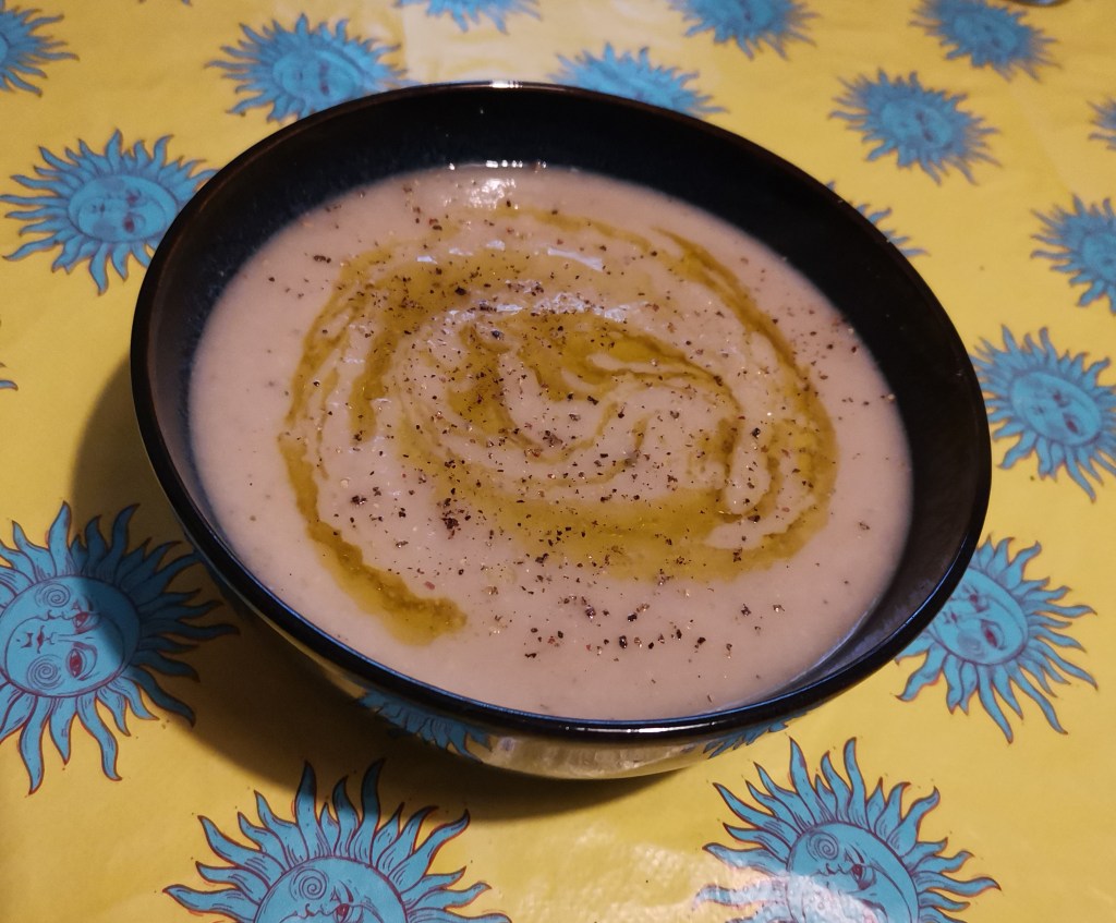 A bowl of soup, drizzled with olive oil and topped with black pepper. The bowl sits on a yellow tablecloth with blue sun faces.